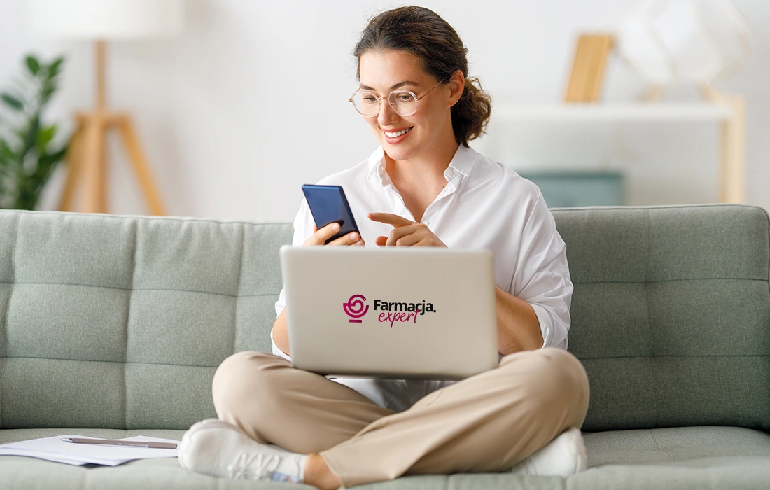 Uśmiechnięta kobieta siedzi ze skrzyżowanymi nogami na sofie, korzystając z telefonu i laptopa. Na laptopie widnieje logo Farmacja expert. Ma na sobie okulary i swobodne ubranie.