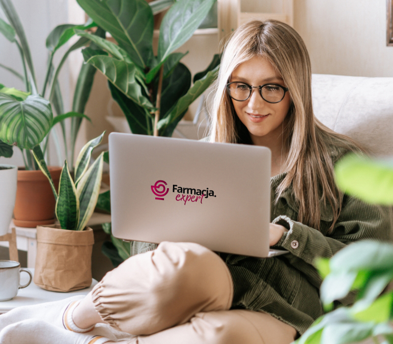 Kobieta w okularach siedzi wśród roślin doniczkowych, korzysta z laptopa z logo Farmacja Expert, wygląda na skupioną i zrelaksowaną w przytulnym, jasnym pokoju.