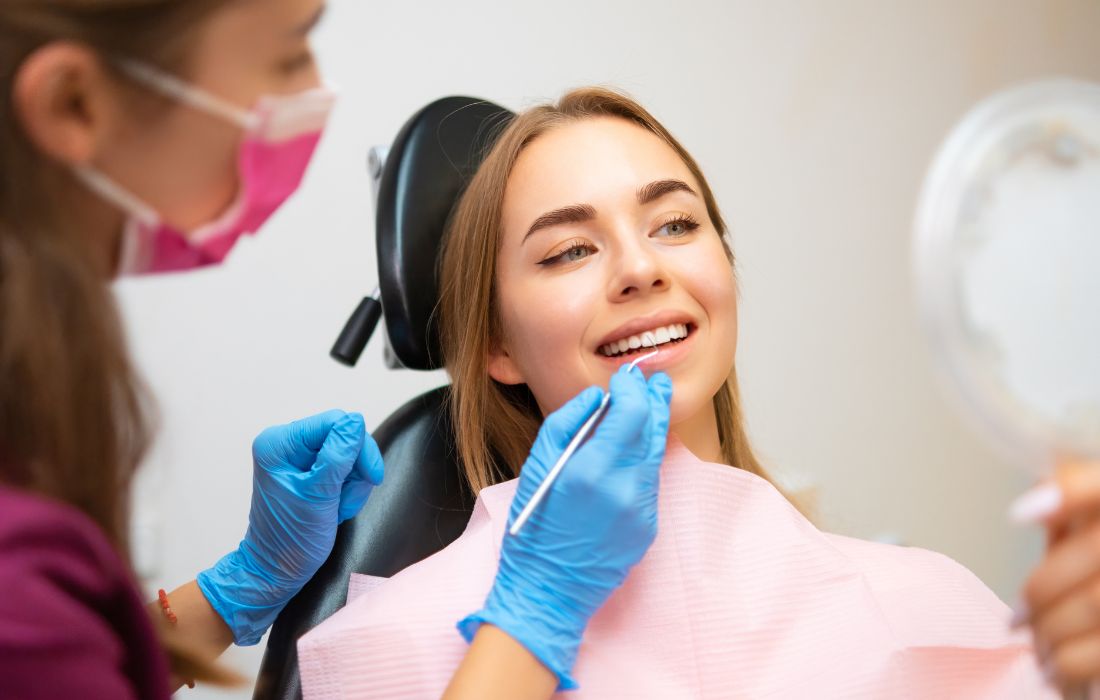 Dentysta w rękawiczkach i masce bada zęby uśmiechniętej młodej kobiety siedzącej na fotelu dentystycznym, trzymającej lusterko.