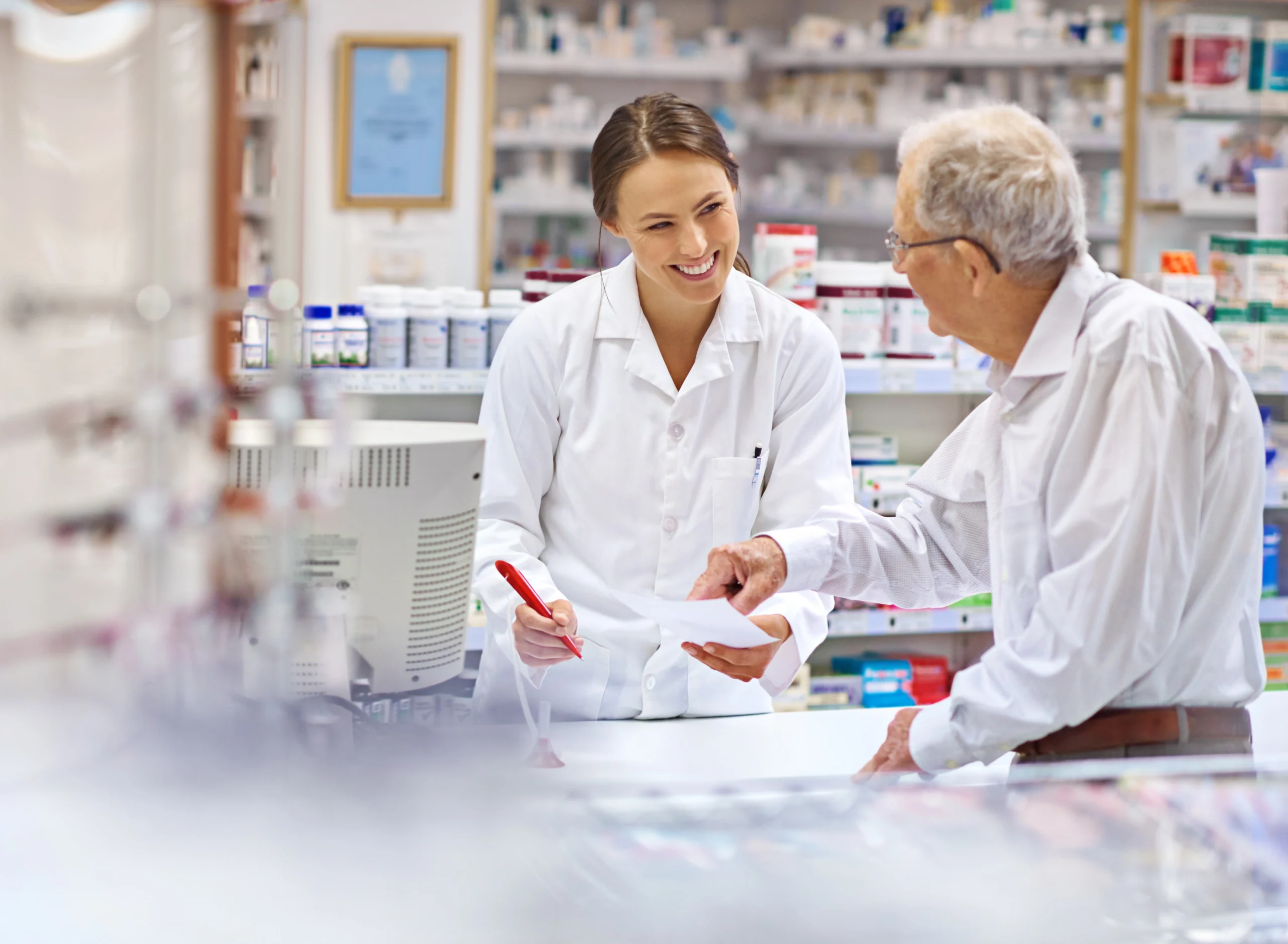 Uśmiechnięta farmaceutka pomaga starszemu klientowi przy ladzie apteki. W tle widać półki z lekami.
