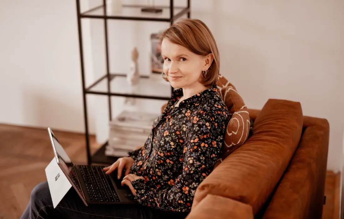 Kobieta w kwiecistej bluzce siedzi na brązowej sofie, trzyma laptopa na kolanach i patrzy w górę na kamerę. W tle widać czarną półkę z dekoracjami i papierami.