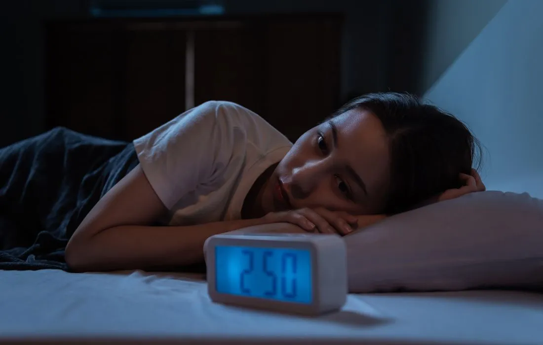 Kobieta leży nocą w łóżku, nie mogąc zasnąć, z głową opartą na poduszce, obok cyfrowego zegara wskazującego godzinę 2:50.