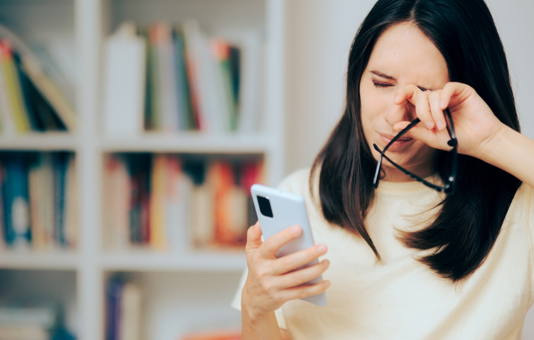 Kobieta stojąca przed regałem z książkami, pociera oczy, trzyma okulary i patrzy w telefon.