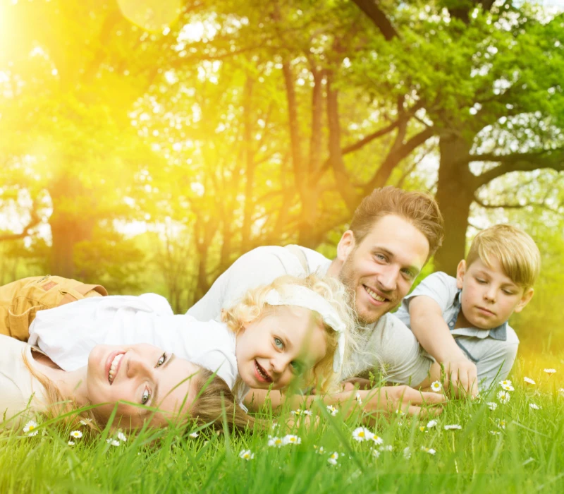 Rodzina składająca się z czterech osób leży na trawie w rozświetlonym słońcem parku, uśmiechając się. Otaczają ich drzewa o zielonych liściach, tworząc ciepłą, radosną atmosferę.
