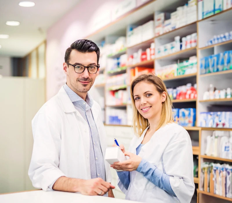 Dwóch farmaceutów w białych kitlach stoi przy ladzie aptecznej, uśmiecha się, w tle widać półki z lekami i produktami medycznymi.