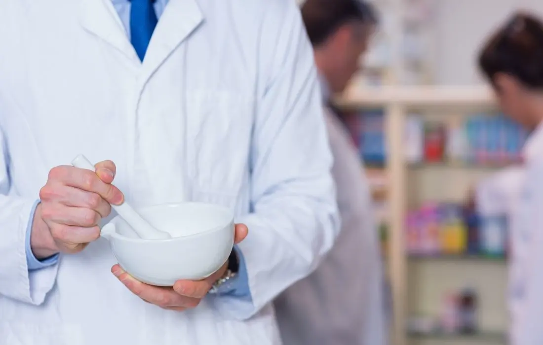 Osoba w białym fartuchu laboratoryjnym używa moździerza i tłuczka, w tle widać półki i dwie rozmazane postacie, prawdopodobnie w aptece lub laboratorium.