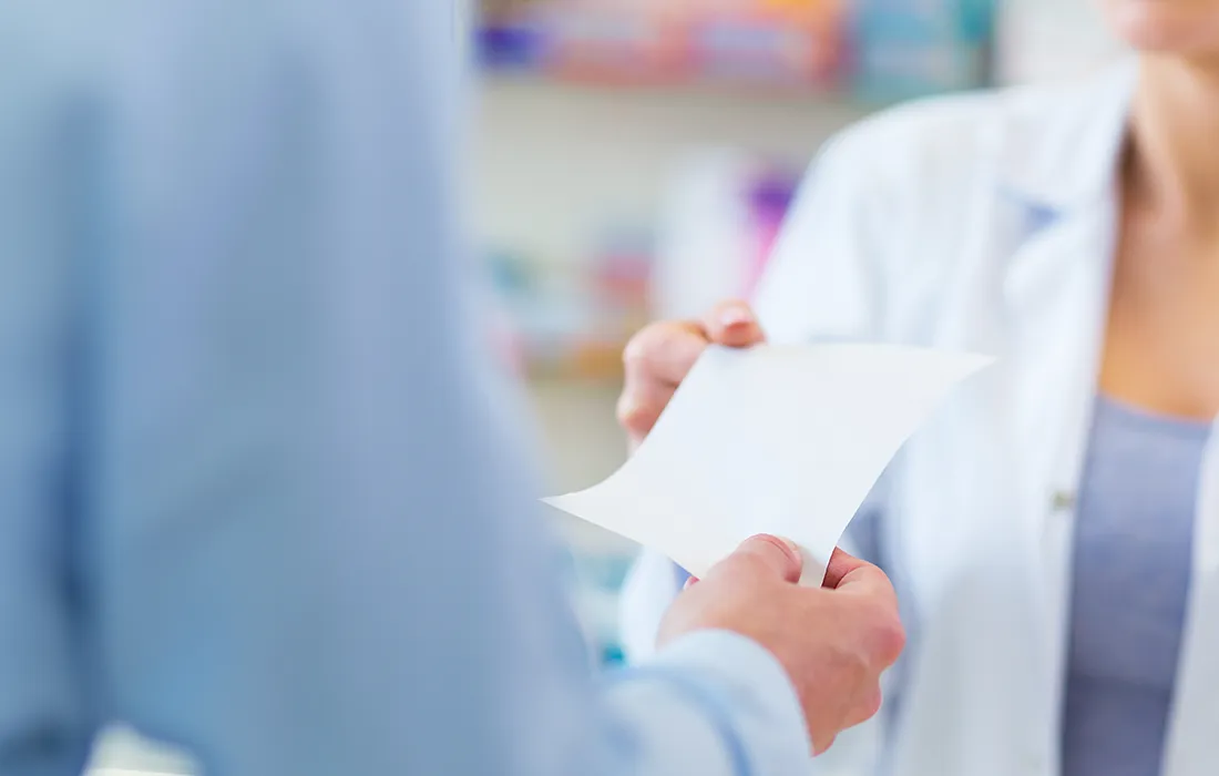 Osoba wręcza receptę lub kartkę papieru farmaceucie lub pracownikowi służby zdrowia w białym fartuchu przy ladzie aptecznej.
