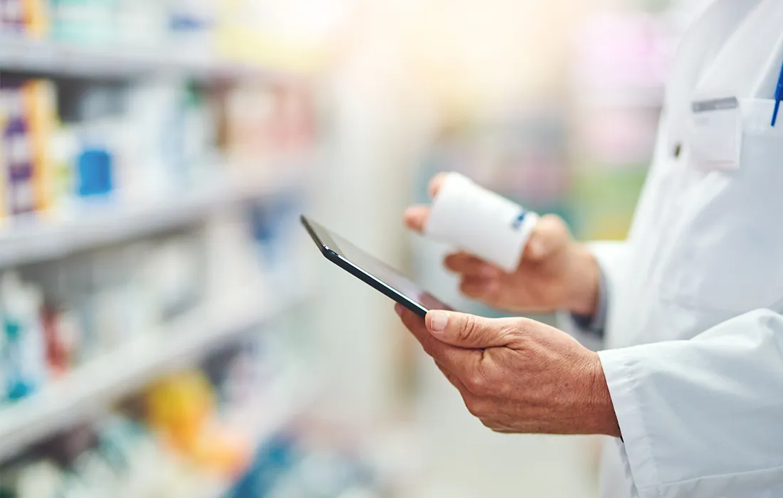 Farmaceuta w białym kitlu trzyma w jednej ręce buteleczkę z lekiem, a w drugiej posługuje się tabletem cyfrowym, stojąc przed półkami z różnymi lekami.