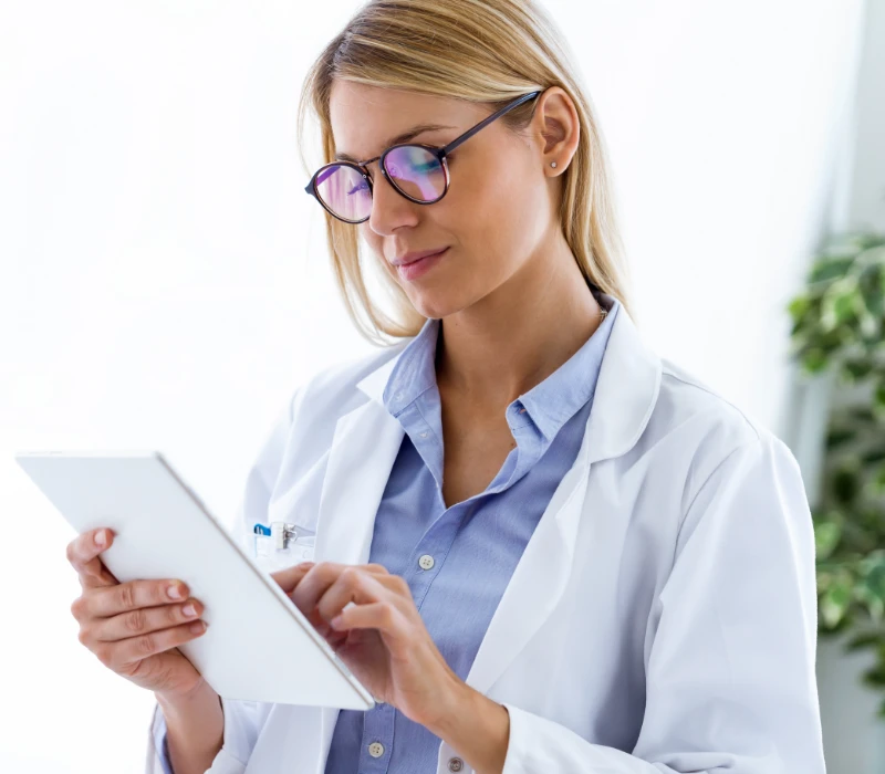 Kobieta w białym fartuchu laboratoryjnym i okularach korzysta z tabletu cyfrowego, stojąc w pomieszczeniu, na tle rozmazanej rośliny.