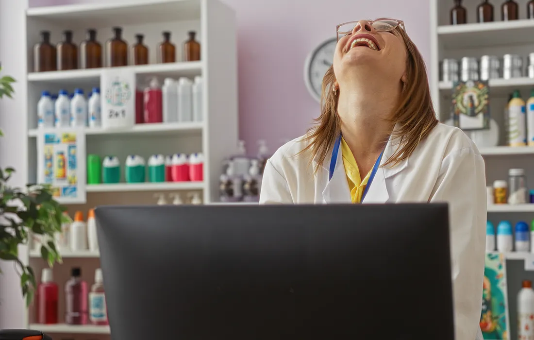 Kobieta w białym fartuchu laboratoryjnym siedzi przy biurku, śmieje się, odchylając głowę do tyłu, w pokoju za nią, gdzie znajdują się półki z butelkami i innymi przyborami.