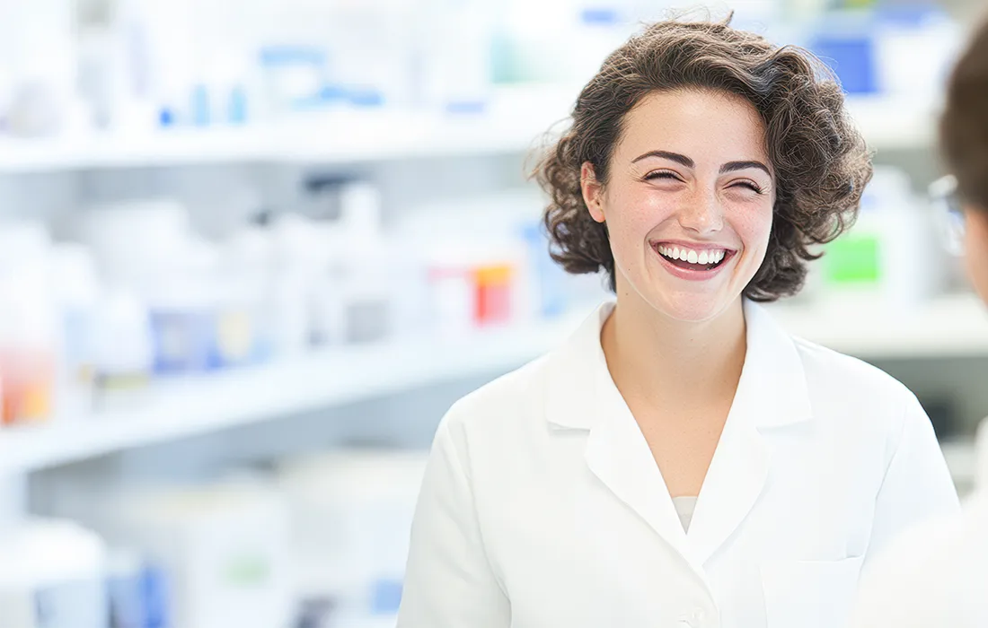 Uśmiechnięta kobieta w białym fartuchu laboratoryjnym stoi w jasnym pomieszczeniu apteki lub laboratorium, w tle rozmywają się półki z butelkami i pojemnikami.
