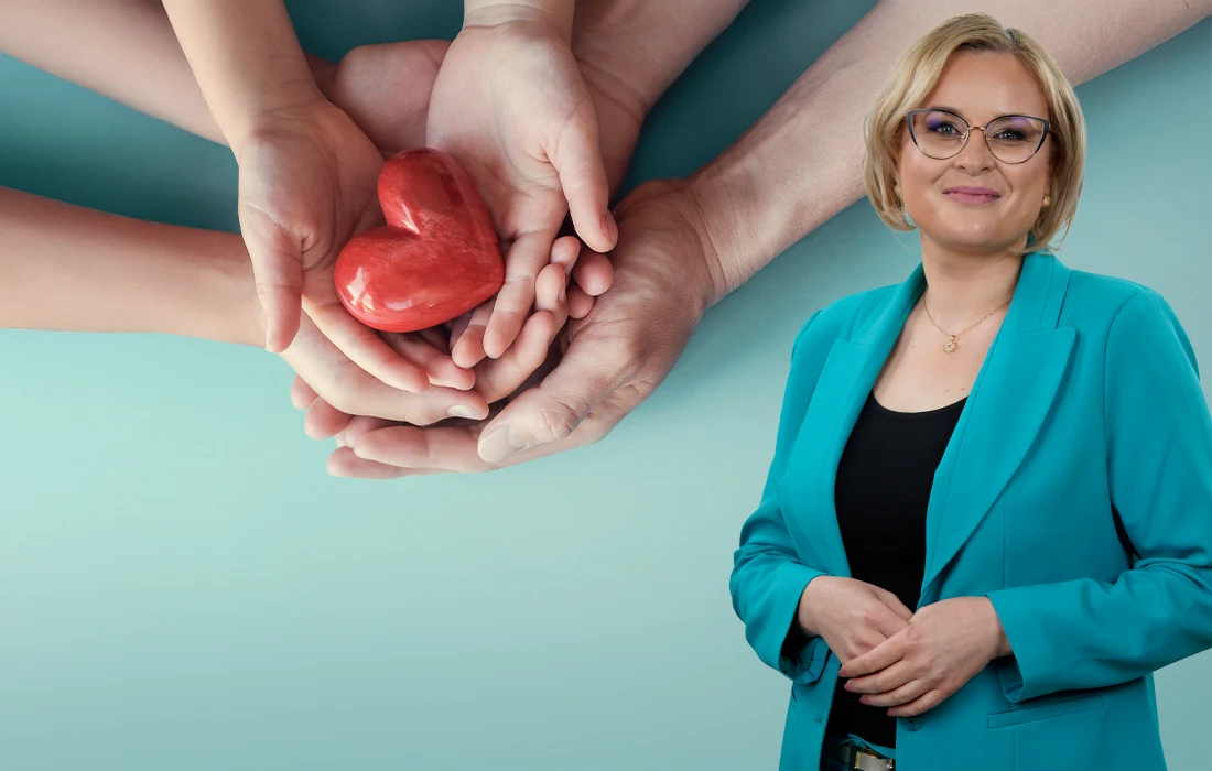 Kobieta w turkusowej marynarce stoi i uśmiecha się, trzymając w dłoniach czerwone serce na niebieskim tle.