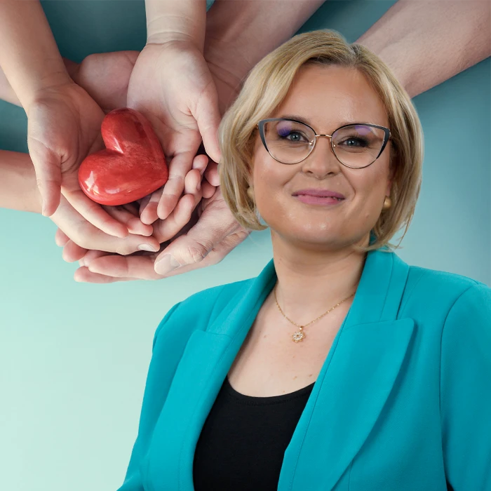 Kobieta w turkusowej marynarce uśmiecha się przed obrazem przedstawiającym dłonie trzymające czerwone serce, symbolizujące troskę i wsparcie.