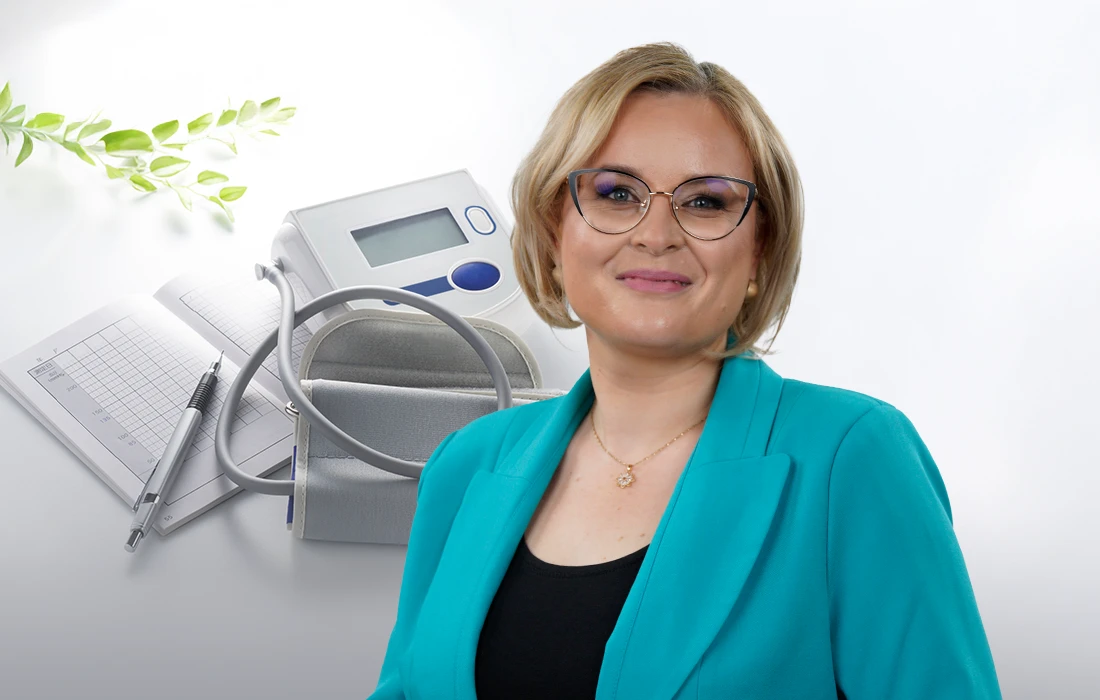 Uśmiechnięta kobieta w okularach i turkusowej marynarce stoi przed cyfrowym ciśnieniomierzem, otwartym notatnikiem i długopisem na jasnym tle.