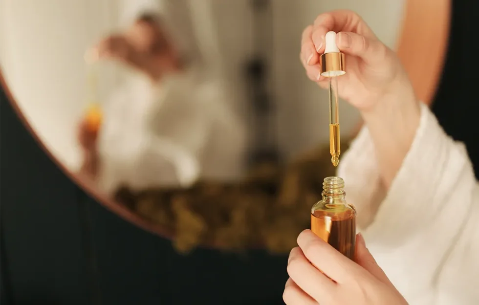 Osoba w szlafroku trzyma zakraplacz nad szklaną butelką olejku, a jej odbicie widoczne jest w okrągłym lustrze w tle.