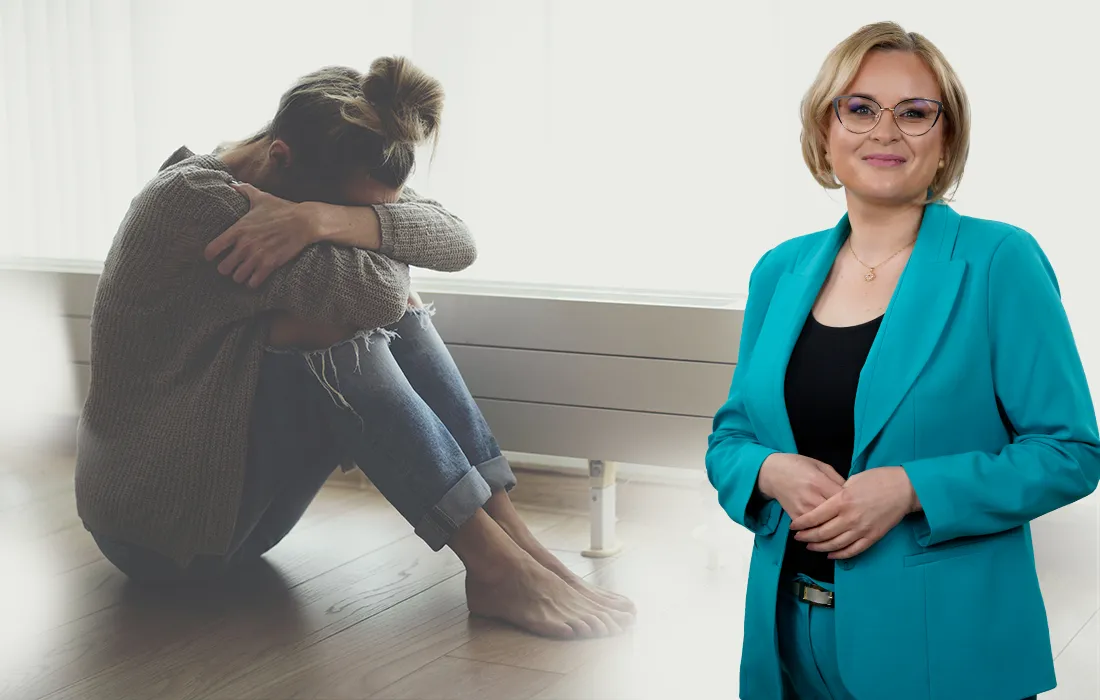Kobieta siedzi na podłodze, obejmując kolana, wyglądając na zrozpaczoną, podczas gdy inna kobieta w turkusowym garniturze uśmiecha się pewnie, stojąc na pierwszym planie.