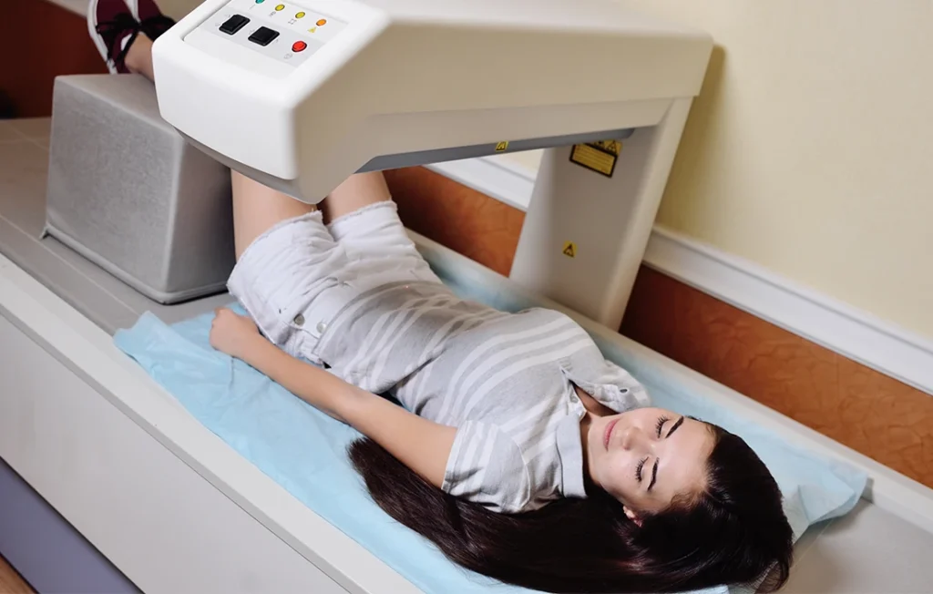 Osoba leży na stole do skanowania medycznego z uniesionymi nogami, poddając się skanowaniu diagnostycznemu, prawdopodobnie badaniu gęstości kości.