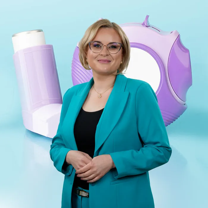 Uśmiechnięta kobieta w turkusowym kostiumie stoi przed powiększonymi zdjęciami fioletowego inhalatora i okrągłego fioletowego urządzenia do leczenia astmy na niebieskim tle.