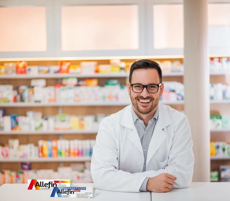 Uśmiechnięty farmaceuta w białym fartuchu siedzi przy ladzie w aptece, z półkami leków za nim i pudełkami produktów Allefin wystawionymi na ladzie.