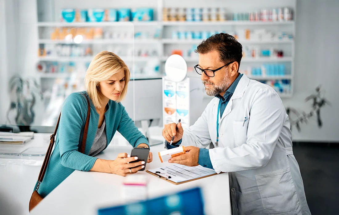 Farmaceuta tłumaczy kobiecie lek przy ladzie aptecznej; kobieta słucha i patrzy na swój telefon, podczas gdy farmaceuta trzyma butelkę z pigułkami i schowek.