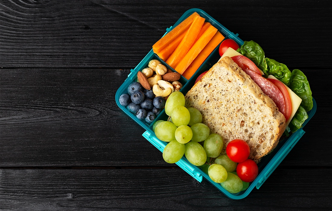 Lunchbox z kanapką, zielonymi winogronami, pomidorkami koktajlowymi, jagodami, orzechami i marchewką na ciemnym drewnianym stole.