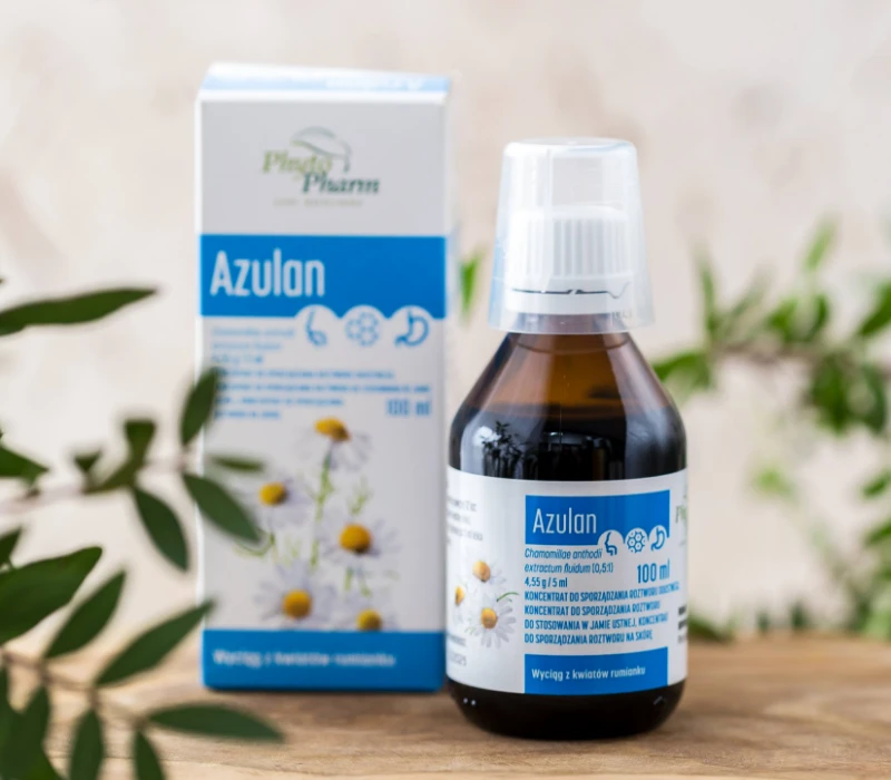 Butelka płynnego ekstraktu ziołowego Azulan stoi przed pudełkiem, oba oznaczone na niebiesko i biało z kwiatami rumianku przedstawionymi na opakowaniu. Zielone liście otaczają scenę.