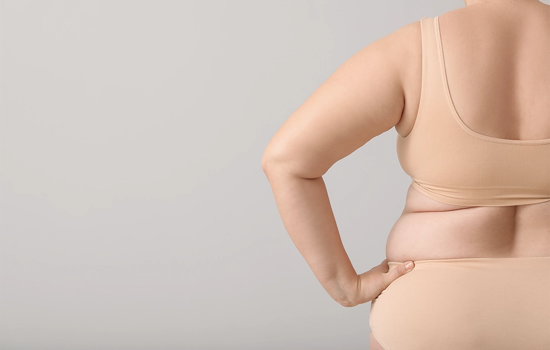 Osoba plus-size ubrana w bieliznę w kolorze nude stoi z jedną ręką na biodrze, odwrócona od kamery na prostym tle.
