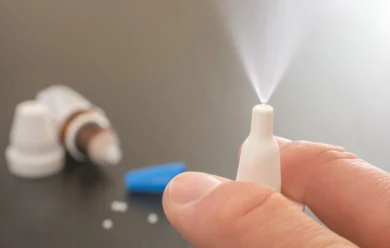 Ręka rozpyla mgiełkę z białej butelki z aerozolem do nosa, z inną butelką i niebieską nasadką rozmytą w tle.