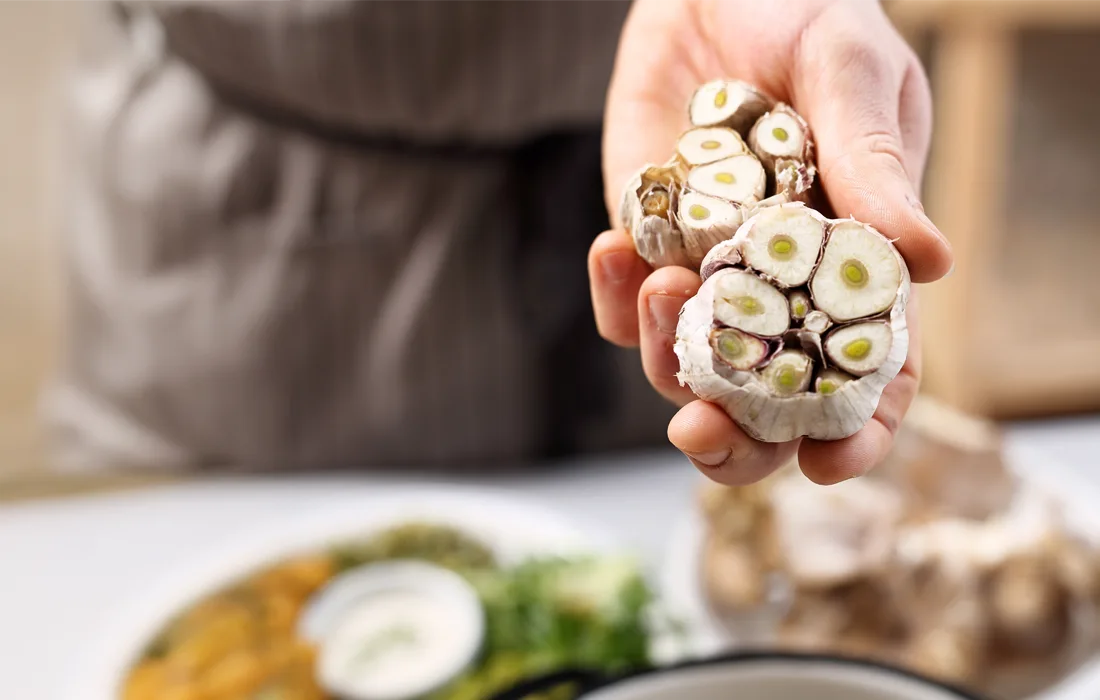 Osoba trzymająca przekrojone na pół główki czosnku, pokazująca przekroje, z rozmytą kuchnią i talerzem z jedzeniem w tle.