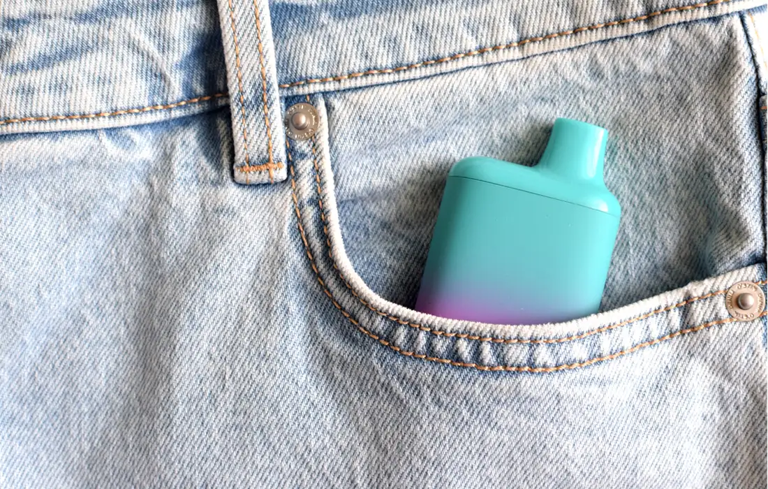 Urządzenie vape w kolorze turkusowym i fioletowym częściowo widoczne w przedniej kieszeni jasnoniebieskich dżinsów.