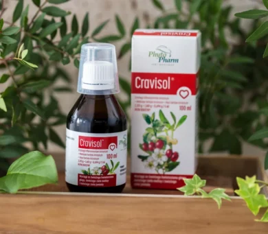 Butelka suplementu w płynie Cravisol z miarką na górze, umieszczona obok pudełka, otoczona zielonymi roślinami na drewnianej powierzchni.