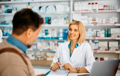 Uśmiechnięty farmaceuta w białym fartuchu rozmawia z klientem po drugiej stronie lady w aptece wypełnionej lekami i produktami zdrowotnymi.