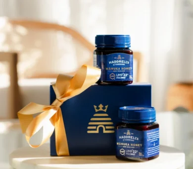 Dwa słoiki miodu Haddrells Manuka w niebieskim pudełku prezentowym przewiązanym złotą wstążką, ustawione w jasnym, delikatnie oświetlonym pomieszczeniu.