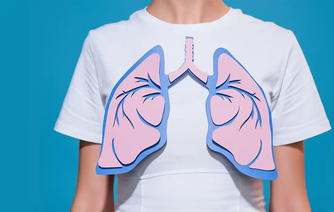 Osoba ubrana w białą koszulkę z wyciętą z papieru ilustracją ludzkich płuc umieszczoną na klatce piersiowej, na niebieskim tle.