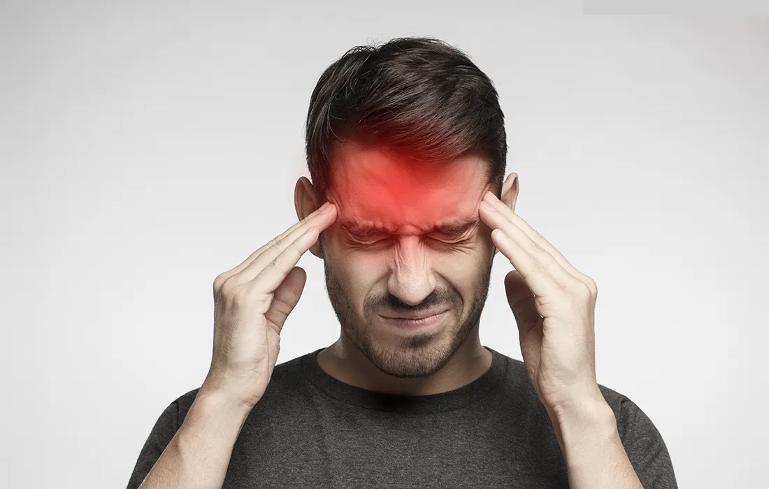Mężczyzna dotyka skroni obiema rękami, z zamkniętymi z bólu oczami i czerwonym obszarem na czole, wskazującym na ból głowy.