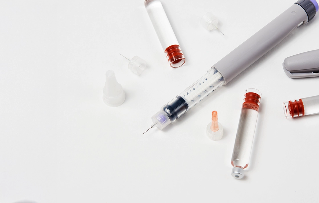Kilka fiolek z czerwonym płynem, igły do strzykawek i pen insulinowy są ułożone na białej powierzchni.