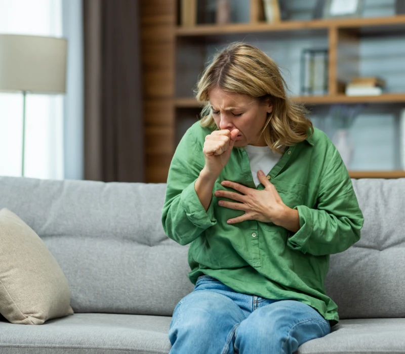 Kobieta w zielonej koszuli siedzi na kanapie, kaszle w pięść i trzyma się za klatkę piersiową, wyglądając na złą.