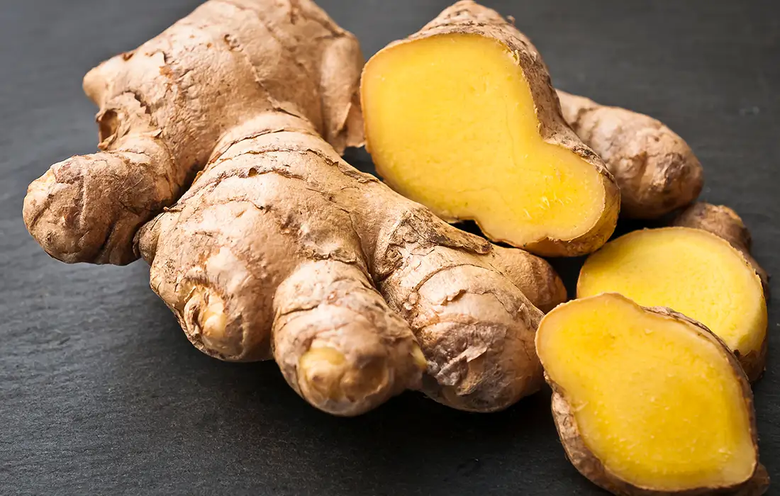 Świeży korzeń imbiru z opaloną skórką, pokazany w całości i z kilkoma plasterkami odsłaniającymi jego żółte wnętrze, na ciemnej powierzchni.