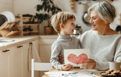 Uśmiechnięta starsza kobieta i młody chłopiec siedzą przy kuchennym stole, trzymając razem rysunek czerwonego serca i patrząc na siebie radośnie.
