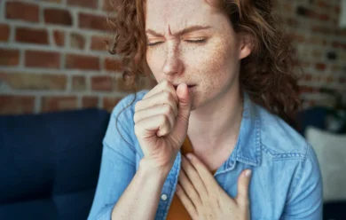 Kobieta z kręconymi włosami i piegami kaszle w rękę, trzymając się za klatkę piersiową, wyglądając niekomfortowo, siedząc w pomieszczeniu z ceglaną ścianą w tle.