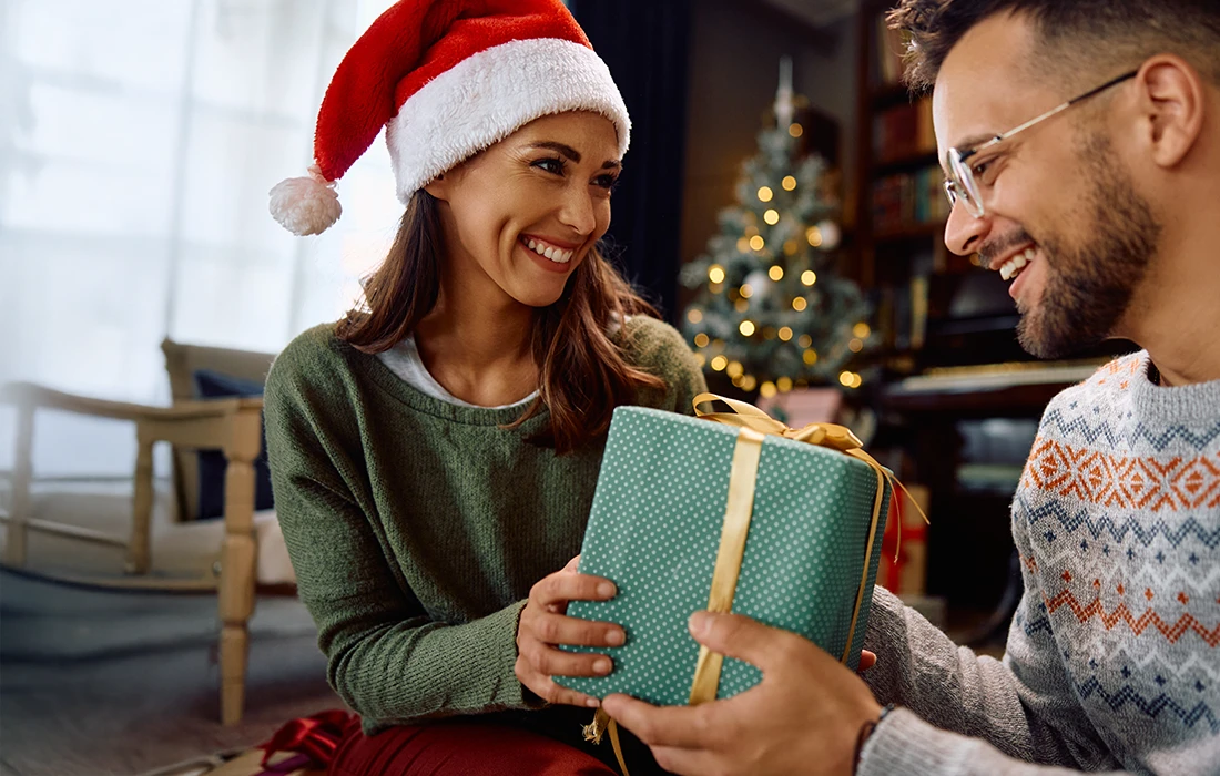 Kobieta w czapce Świętego Mikołaja uśmiecha się, gdy otrzymuje prezent od mężczyzny w świątecznym swetrze, z udekorowaną choinką w tle.