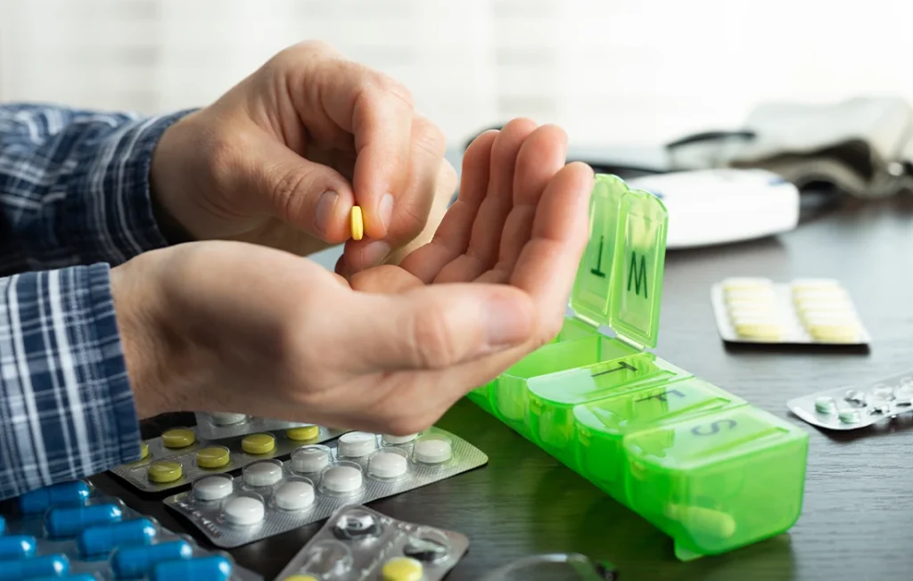 Osoba wkłada żółte tabletki do tygodniowego organizera na tabletki, a na stole leżą różne blistry z lekami.
