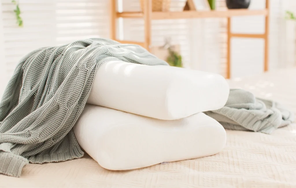 Dwie białe poduszki z pianki memory ułożone na łóżku z jasnoszarym dzianinowym kocem; miękkie naturalne oświetlenie z rozmytymi półkami w tle.