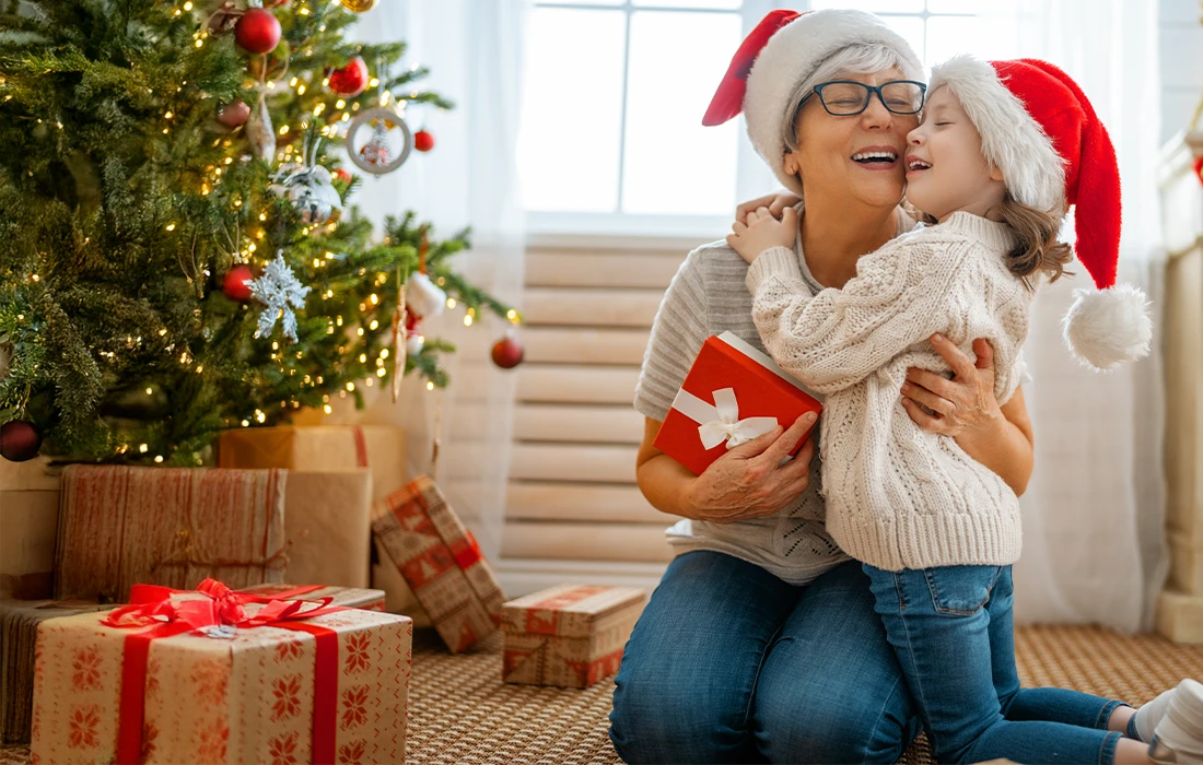 Uśmiechnięta kobieta i dziecko w czapkach Mikołaja przytulają się do choinki otoczonej zapakowanymi prezentami. Kobieta trzyma czerwony prezent z białą wstążką.