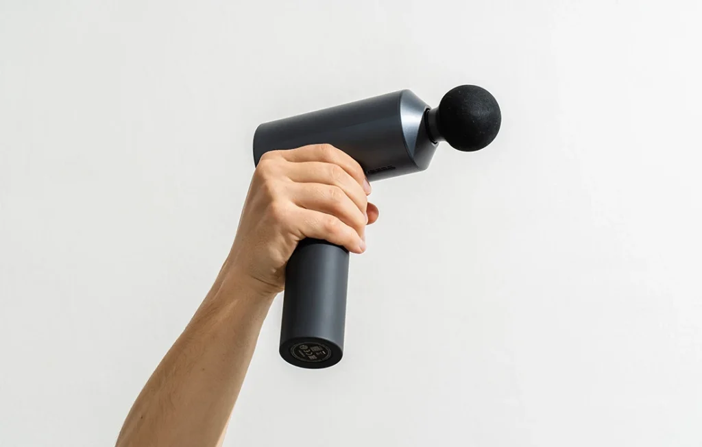 Ręka trzymająca czarny pistolet do masażu z okrągłą nasadką na białym tle.