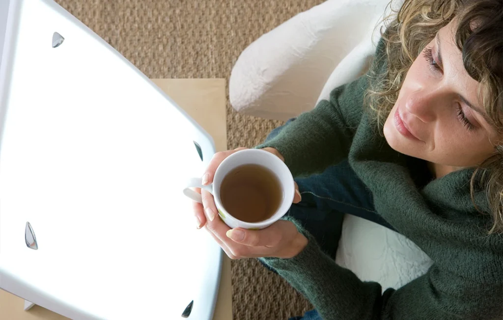 Kobieta trzymająca kubek siedzi przed jasnym pudełkiem do terapii światłem, z zamkniętymi oczami, wyglądając na zrelaksowaną na beżowej podłodze wyłożonej dywanem.