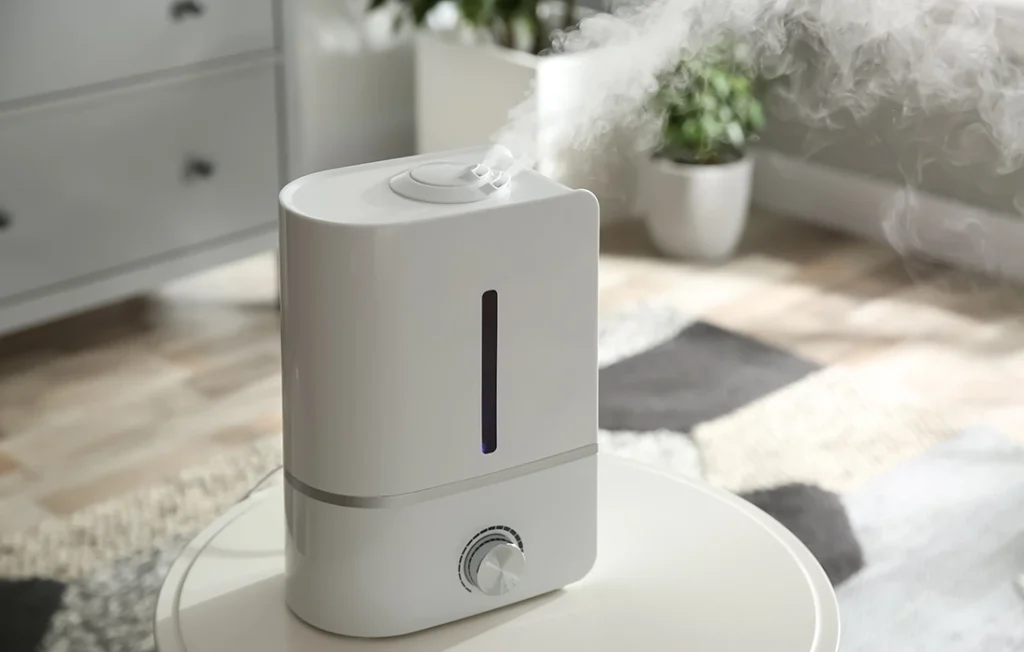 Biały nawilżacz uwalniający mgiełkę stoi na okrągłym stole w jasnym pomieszczeniu z roślinami i nowoczesnym wystrojem w tle.