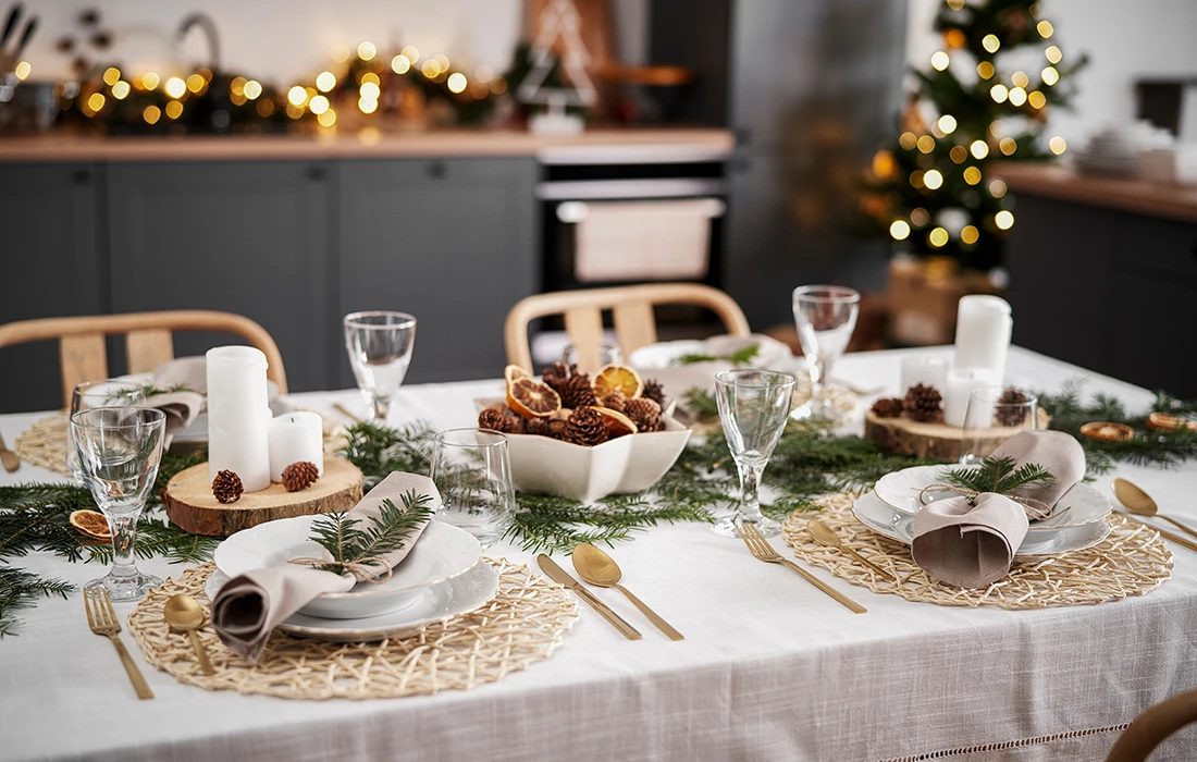 Świąteczny stół zastawiony talerzami, serwetkami, złotymi sztućcami, świecami, szyszkami, zielenią i świątecznymi dekoracjami, z choinką i światełkami w tle.