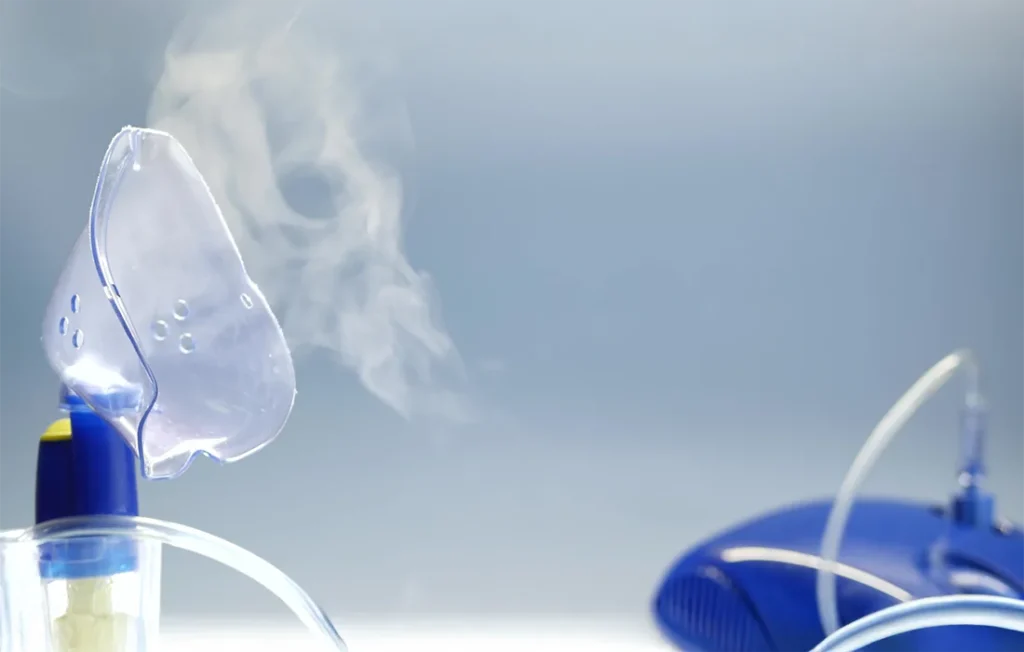 Maska nebulizatora uwalniająca widoczną mgiełkę, z przewodem podłączonym do niebieskiego nebulizatora na jasnoniebieskim tle.