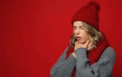 Kobieta w czerwonej czapce i szaliku trzyma się za gardło i wydaje się cierpieć, stojąc przed jednolitym czerwonym tłem.